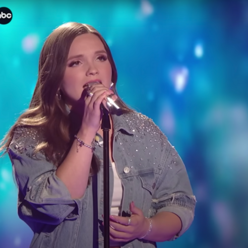 Megan Danielle wearing a blue denim jacket with rhinestones on the shoulders and standing on the American Idol stage against a turquoise background as she sings into a microphone