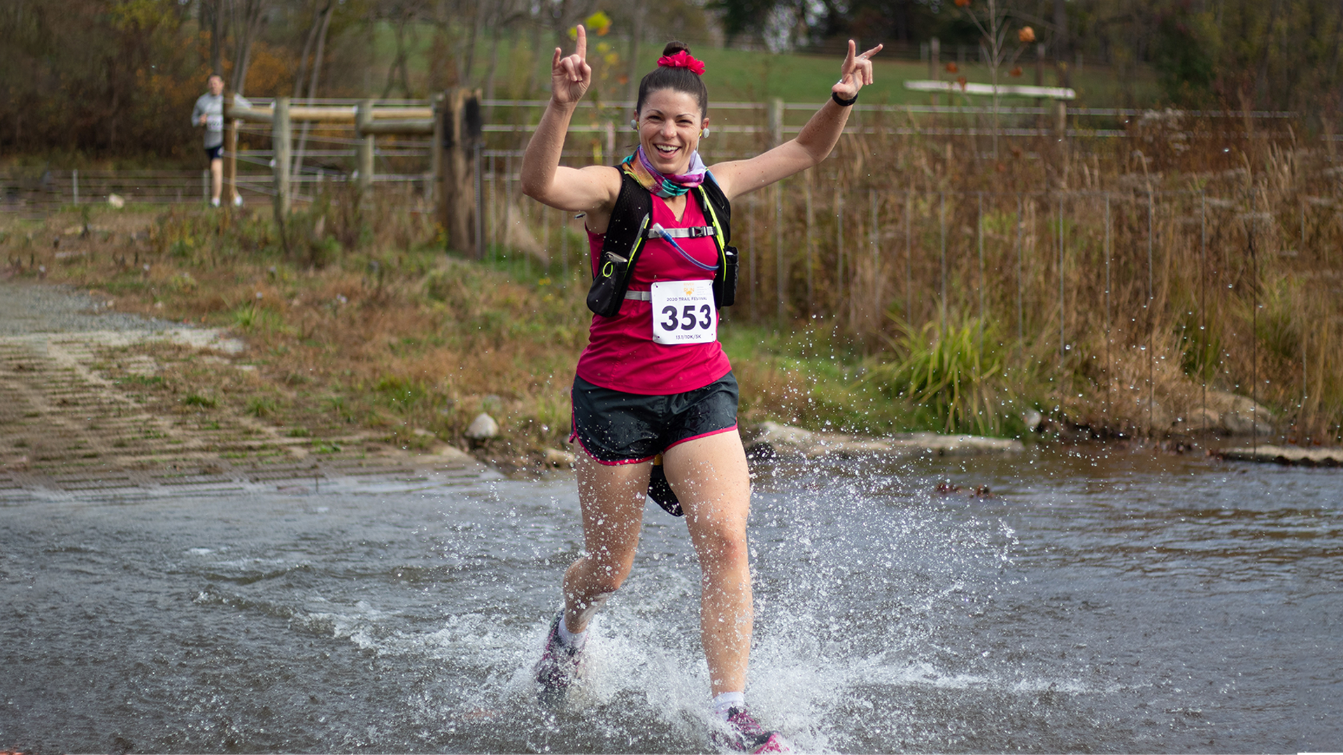 Woman running through a puddle of water while smiling with her hands in the air during the River Valley Trail Race
