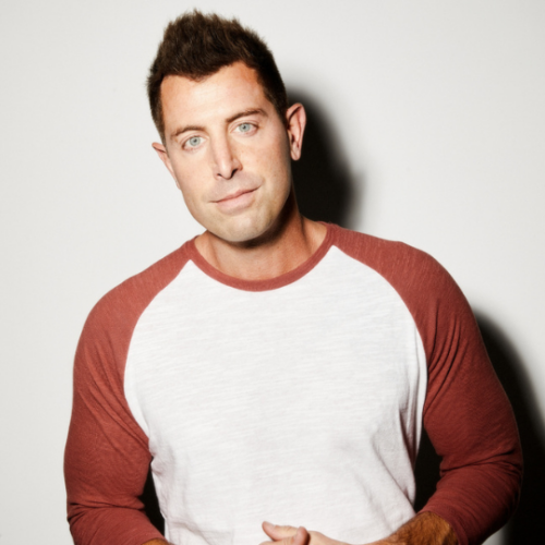 Jeremy Camp wearing a white shirt with burnt orange sleeves and standing against a white background as he smiles softly at the camera