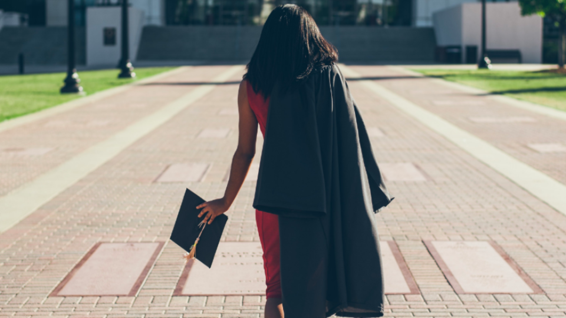 girl wearing a red dress and her black graduation gown draped over her right shoulder as she walks away from the camera while holding her graduation cap in her left hand