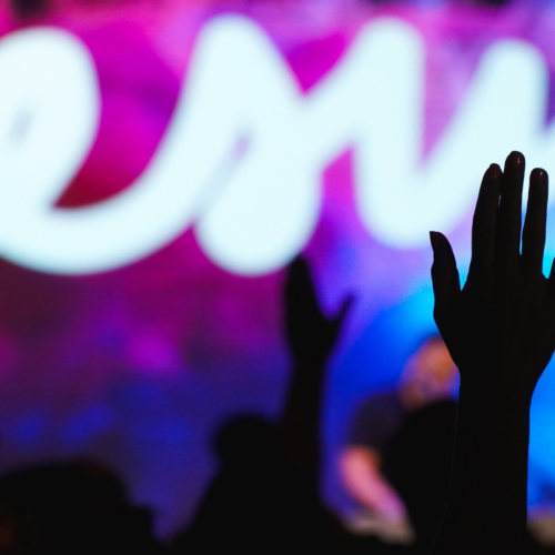 the silhouette of a hand raised in worship during a concert with a screen displaying "Jesus" in the background