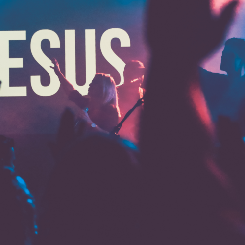 People standing at a worship event with their hands raised up in praise and the name of "Jesus" displayed on the screen on stage