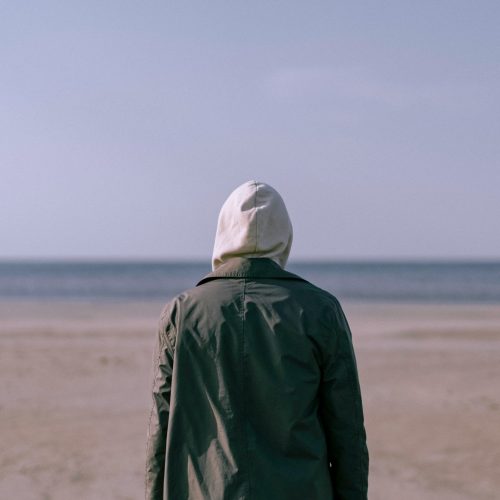 Back of a person with a green jacket looking at the sea on a beach.